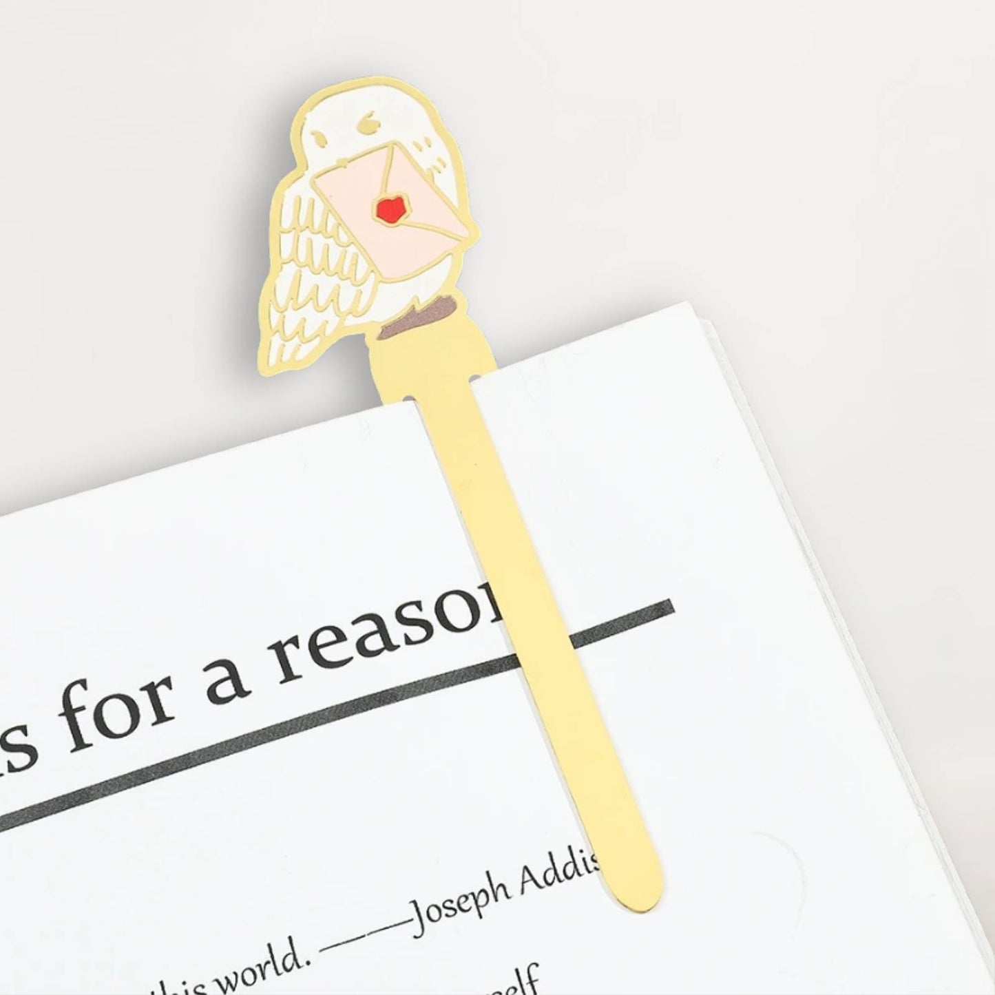 Un marque-page en métal doré représentant un oiseau tenant une lettre rouge, posé sur une page blanche