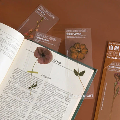 Trois marque-pages en PVC avec des fleurs séchées, posés sur une page d'un livre ouvert sur un fond marron